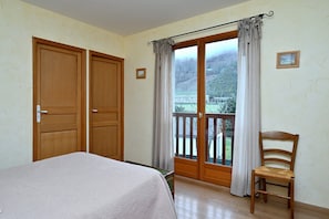 A bedroom with a bed, a wooden chair, a balcony with curtains, and a view of a mountainous landscape.