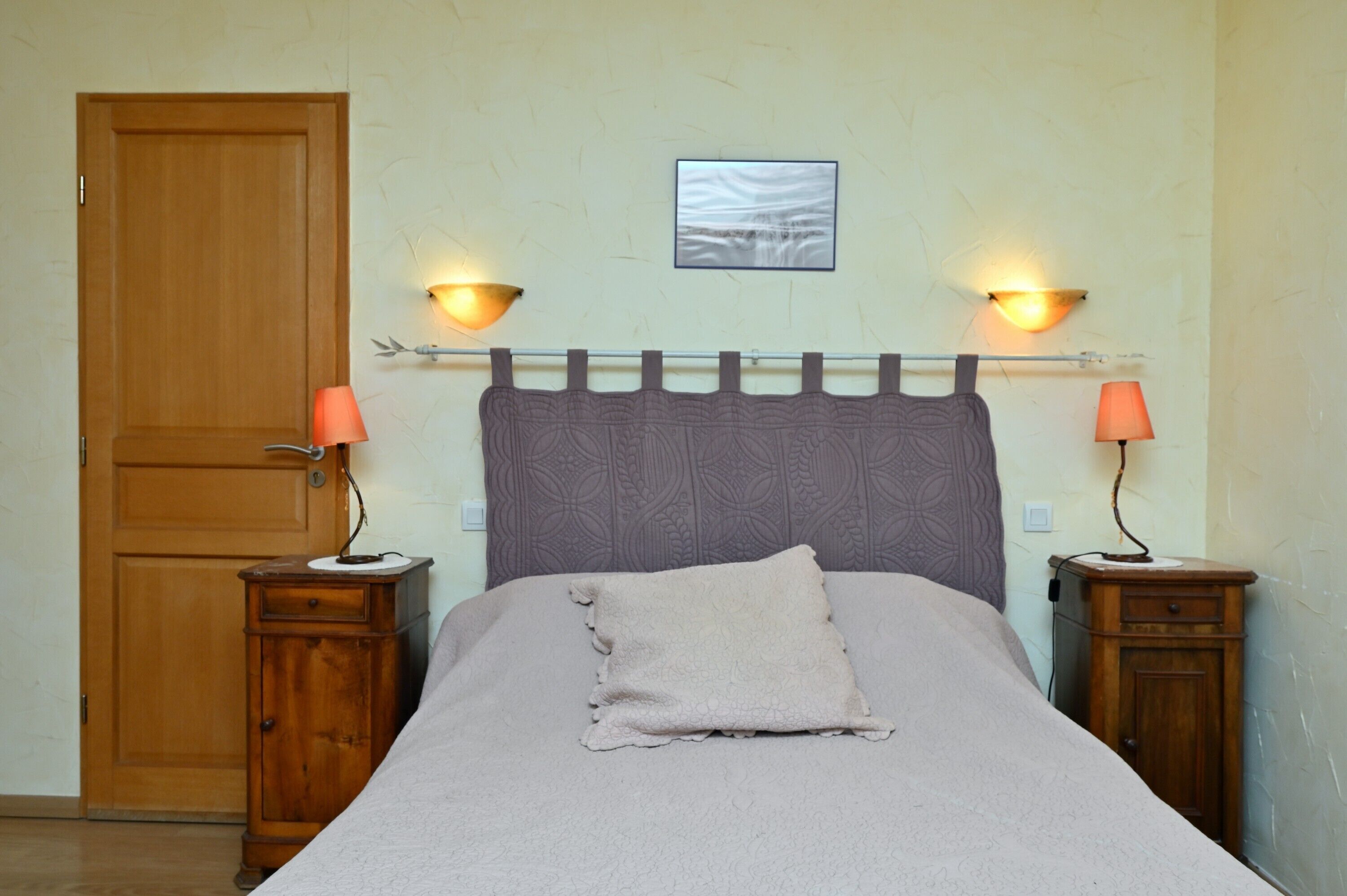 Quarto com porta de madeira, duas mesas de cabeceira com luminárias, uma cama com cabeceira e um quadro na parede.