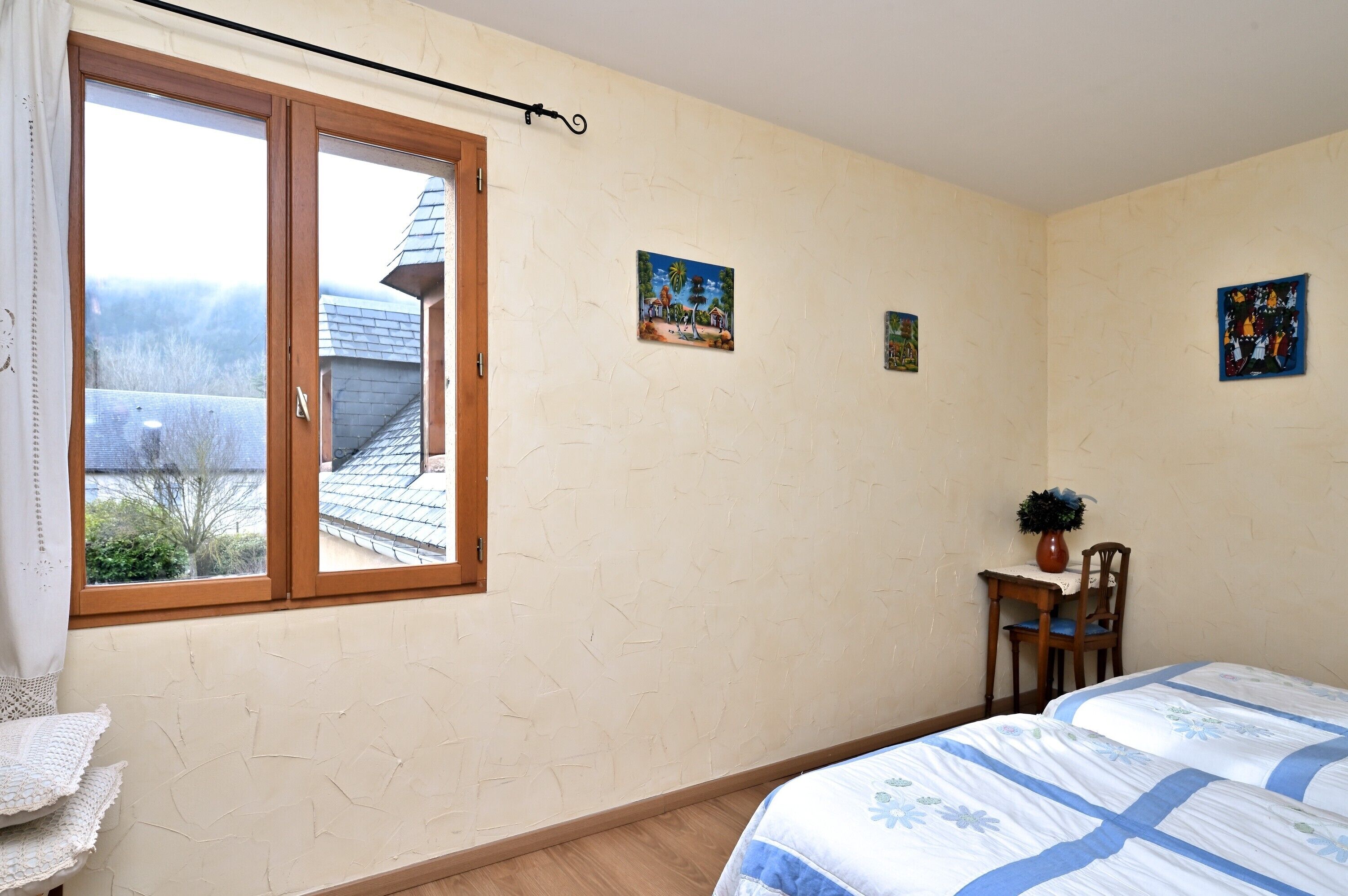 A bedroom with a bed, a desk with a chair, a window with curtains, and a wall with three pictures.
