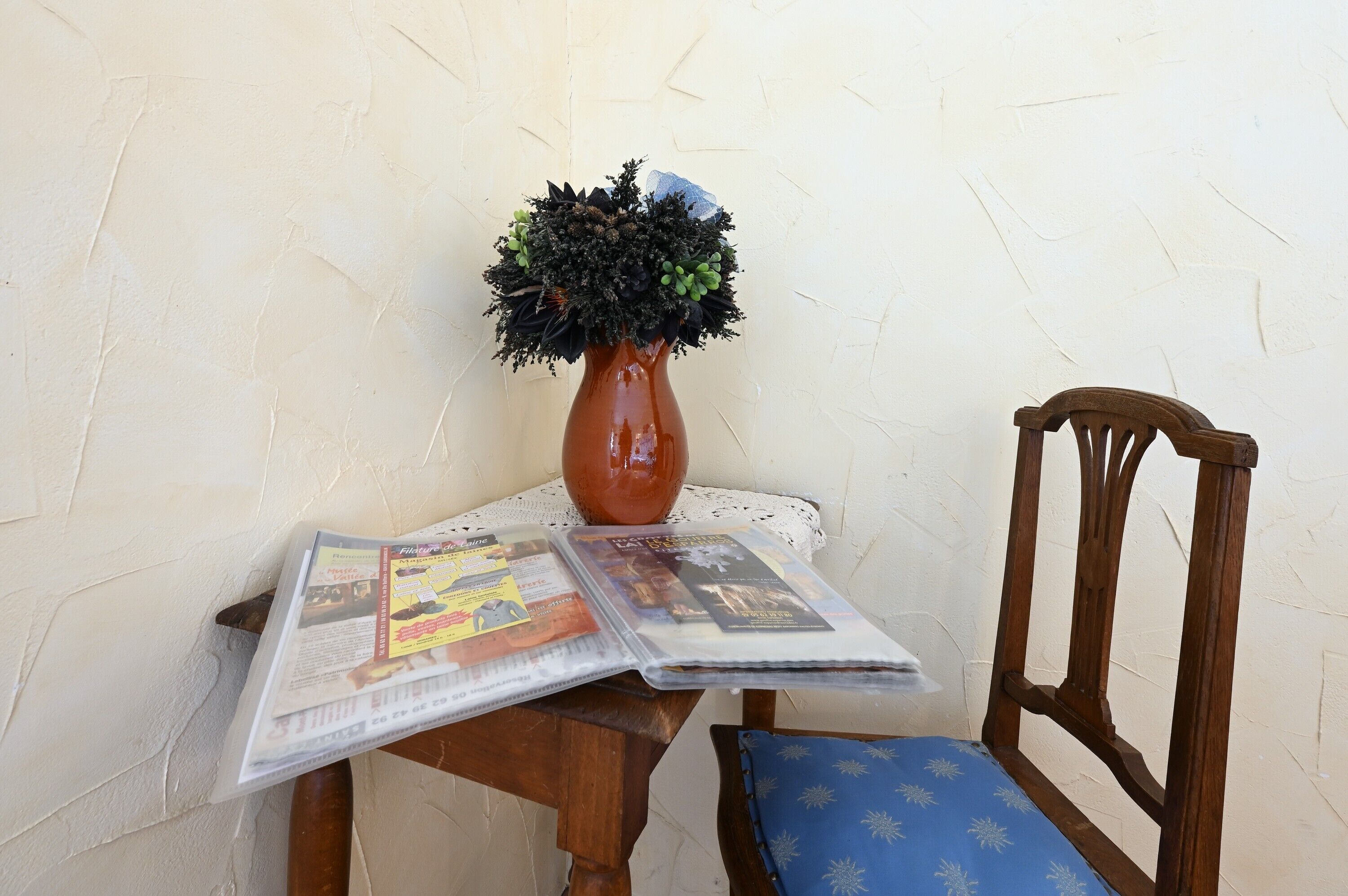 A wooden chair with a blue cushion, a brown vase with a plant, and a newspaper on a table against a textured wall.