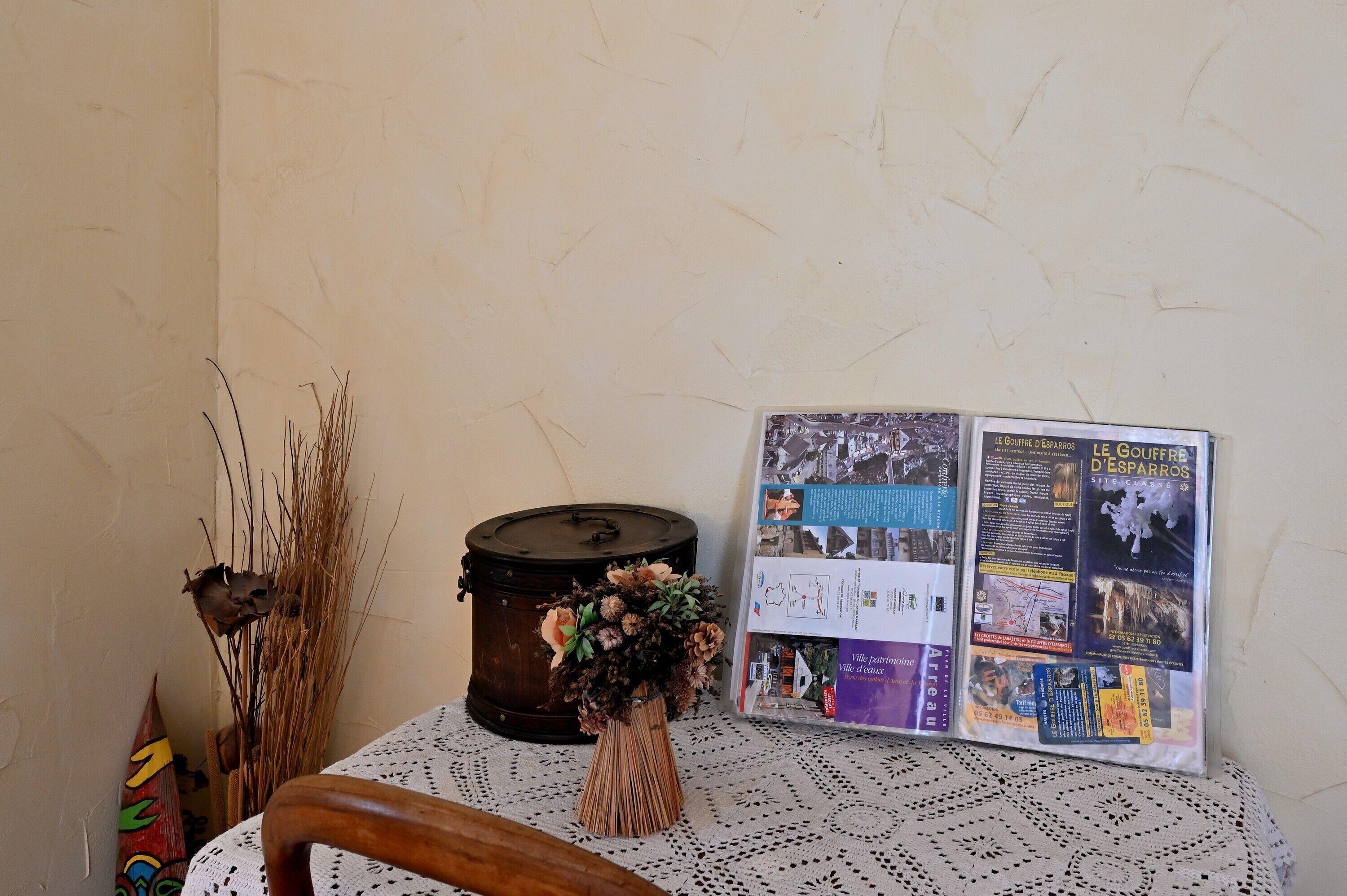 Une table recouverte d’une nappe en dentelle, un vase décoratif contenant des plantes séchées, un tonneau en bois et une brochure.