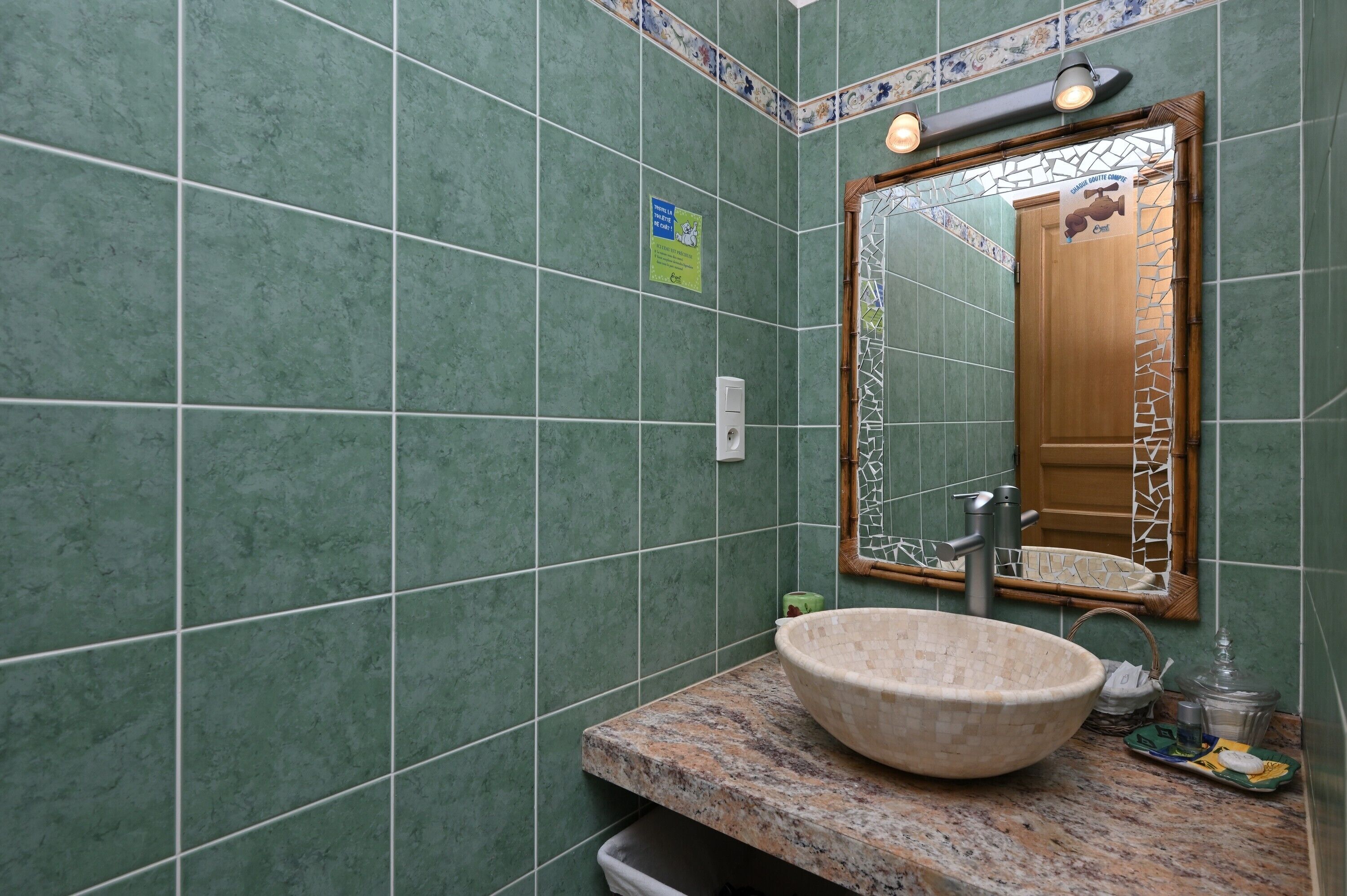 A bathroom with green tiled walls, a large mirror, a granite countertop, and a vessel sink.
