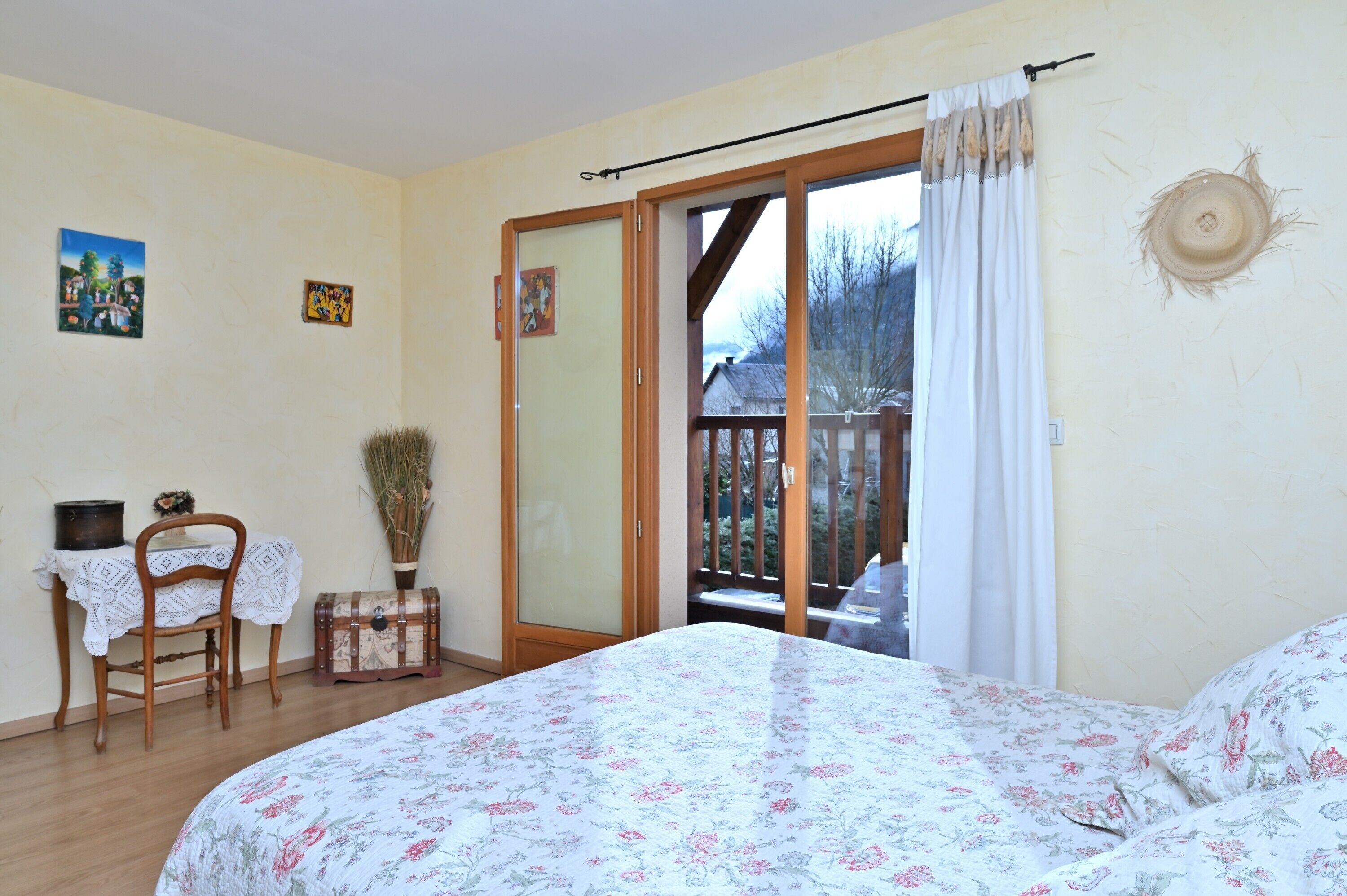 Un dormitorio con cama, una silla, una mesita, un florero y una puerta corrediza de vidrio que da a un balcón.