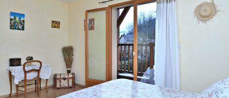 Un dormitorio con cama, una silla, una mesita, un florero y una puerta corrediza de vidrio que da a un balcón.