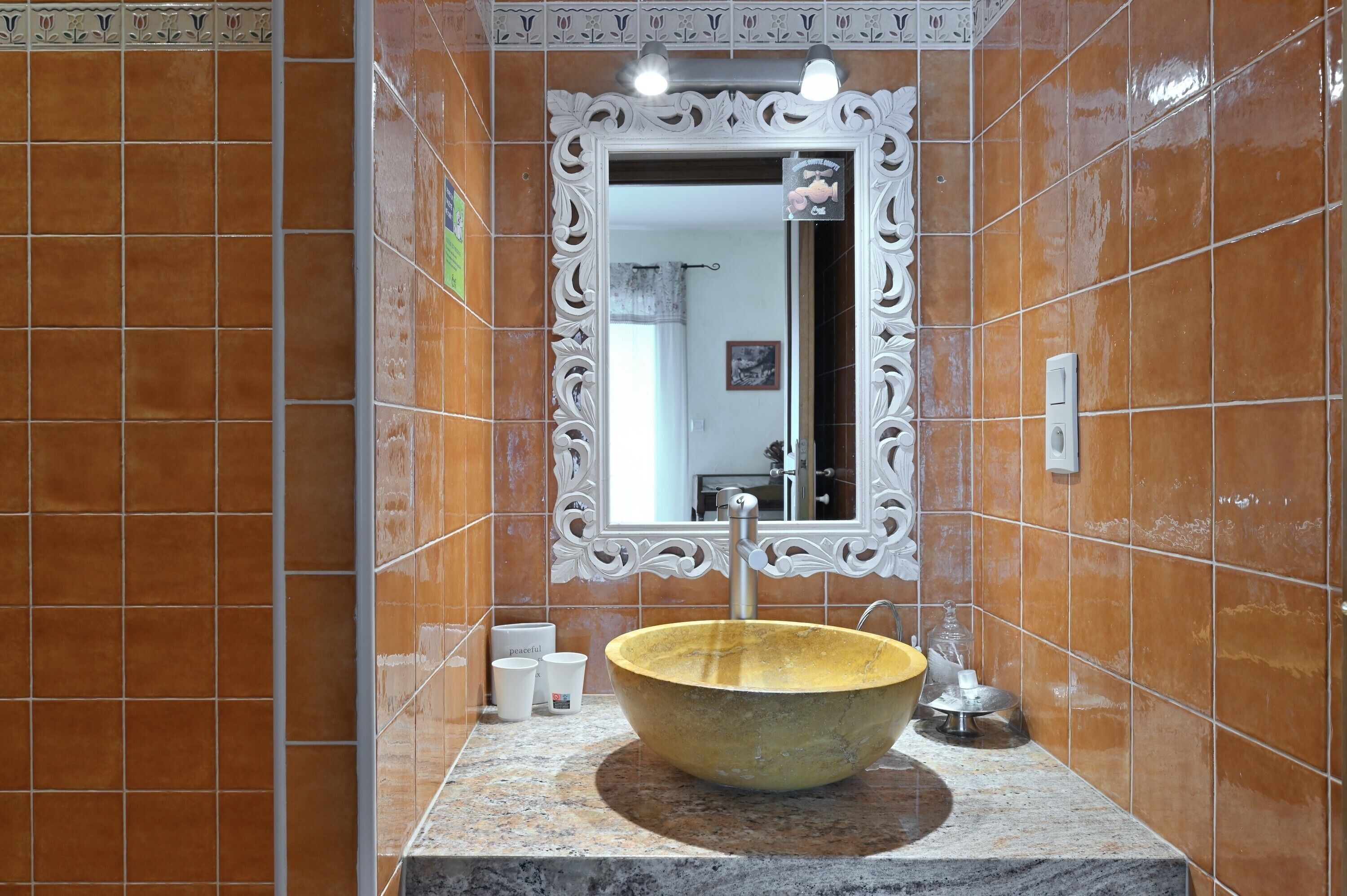 Ein Badezimmer mit einem großen, ovalen Waschbecken, einem Spiegel mit aufwendigem Rahmen und orangefarbenen Fliesenwänden.