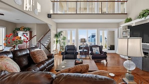 A spacious living room with a high ceiling, a staircase leading to an upper level, and a large window offering a city view. - Beach Diamond - Gulf views, steps to the beach! (Galveston)