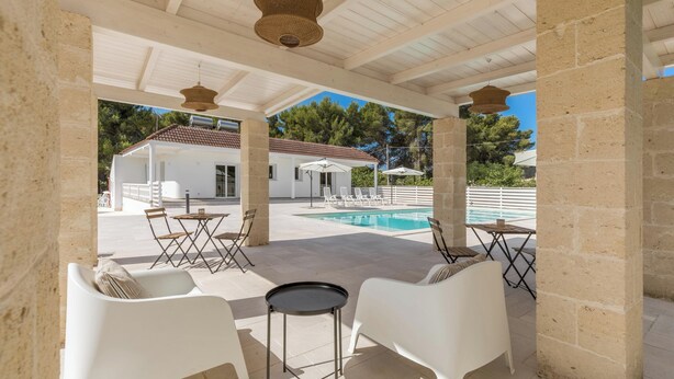Un espacio al aire libre techado con mobiliario blanco, una piscina y un edificio al fondo.