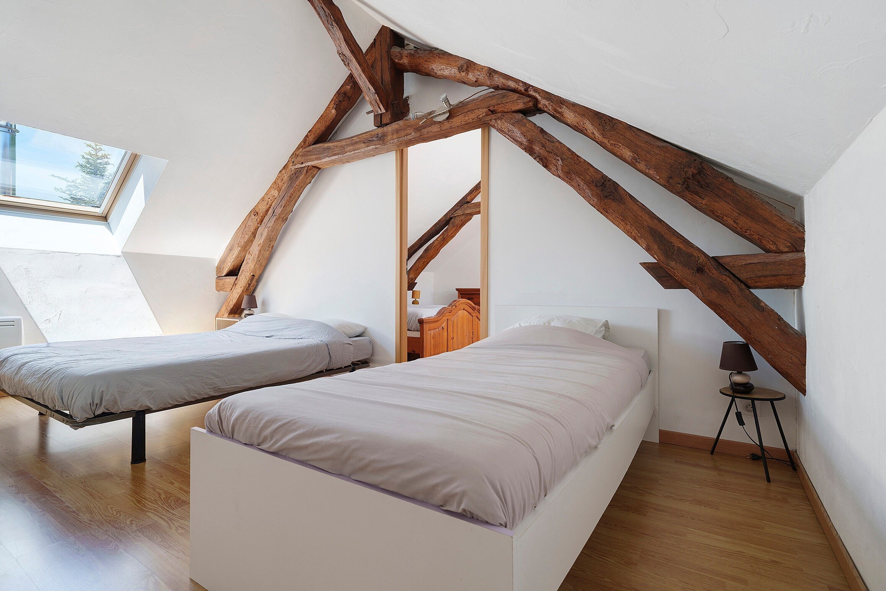 A bedroom with two beds, wooden beams, a skylight, and a small table with a lamp.