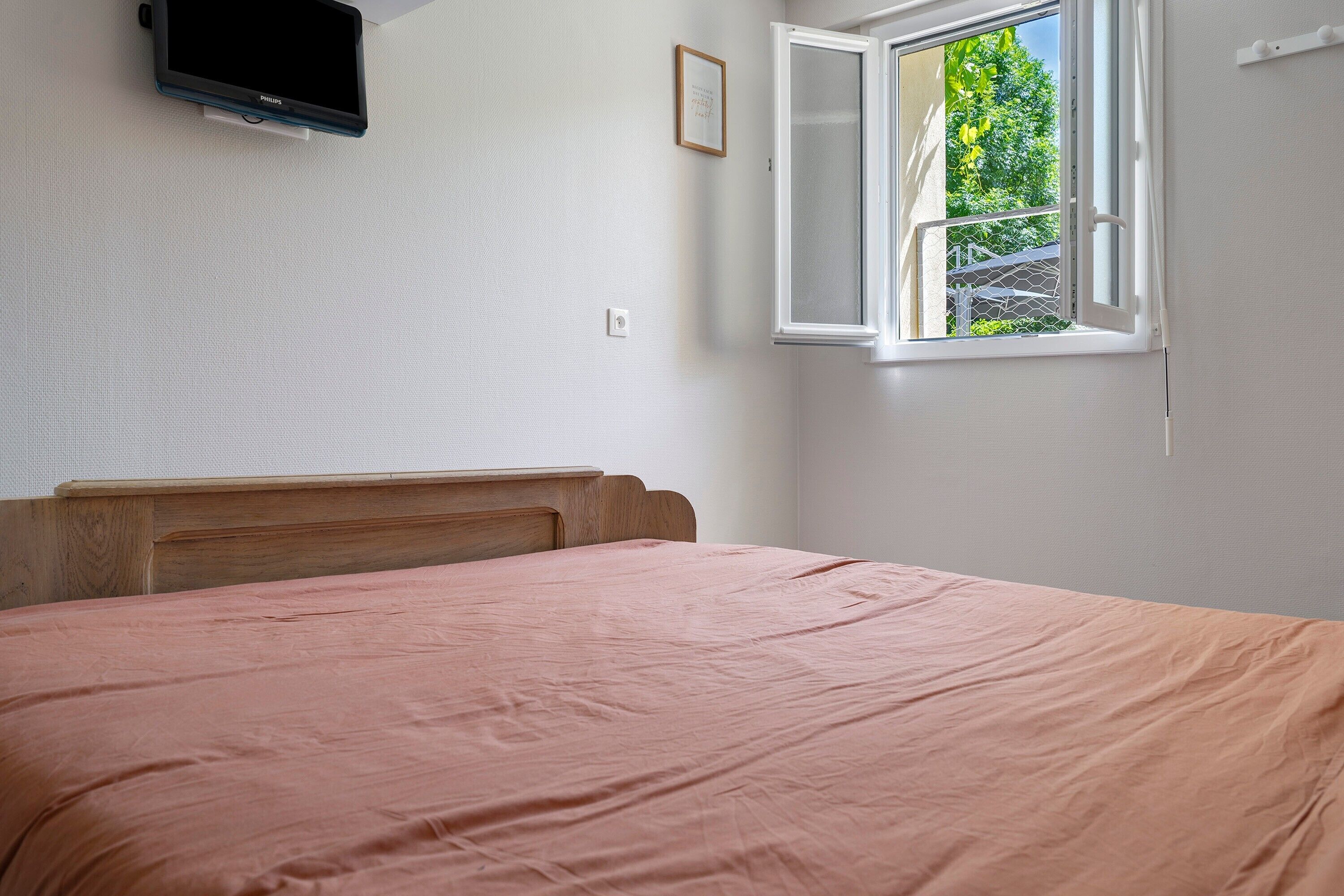 A bedroom with a bed, a mounted TV, a window with an open view, and a door.