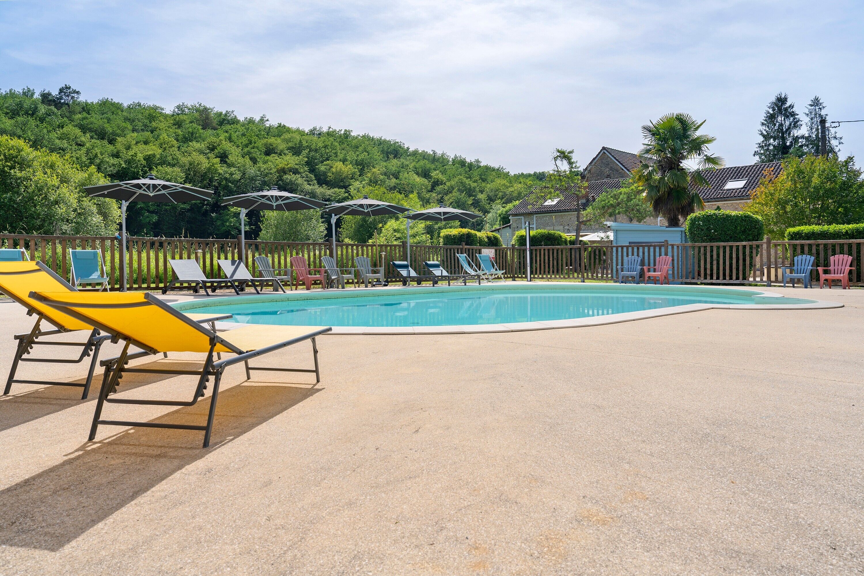 Une piscine entourée d’une clôture en bois et de verdure, avec des chaises longues et des parasols.