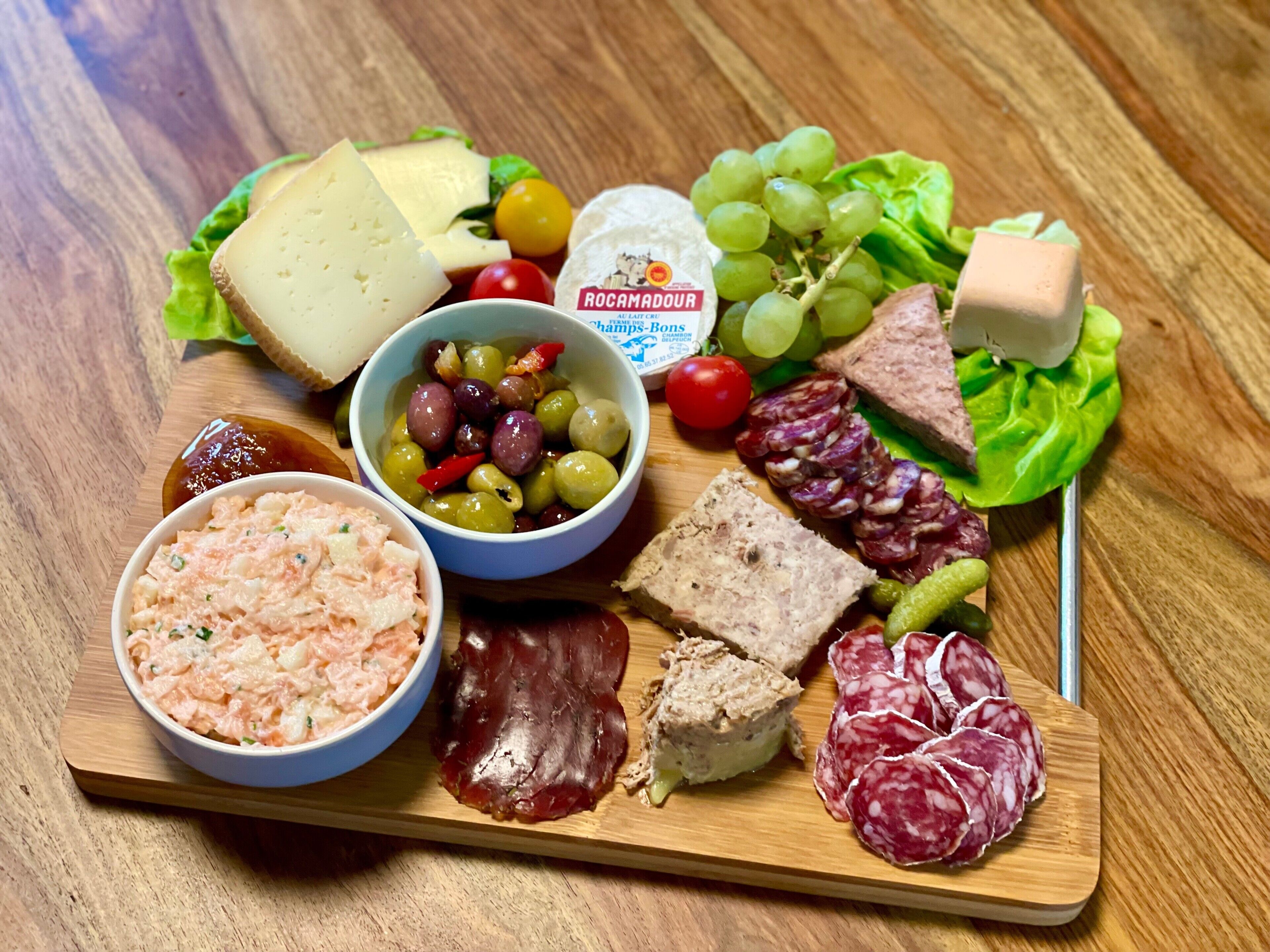 A wooden board with various food items including cheese, olives, grapes, and cured meats.
