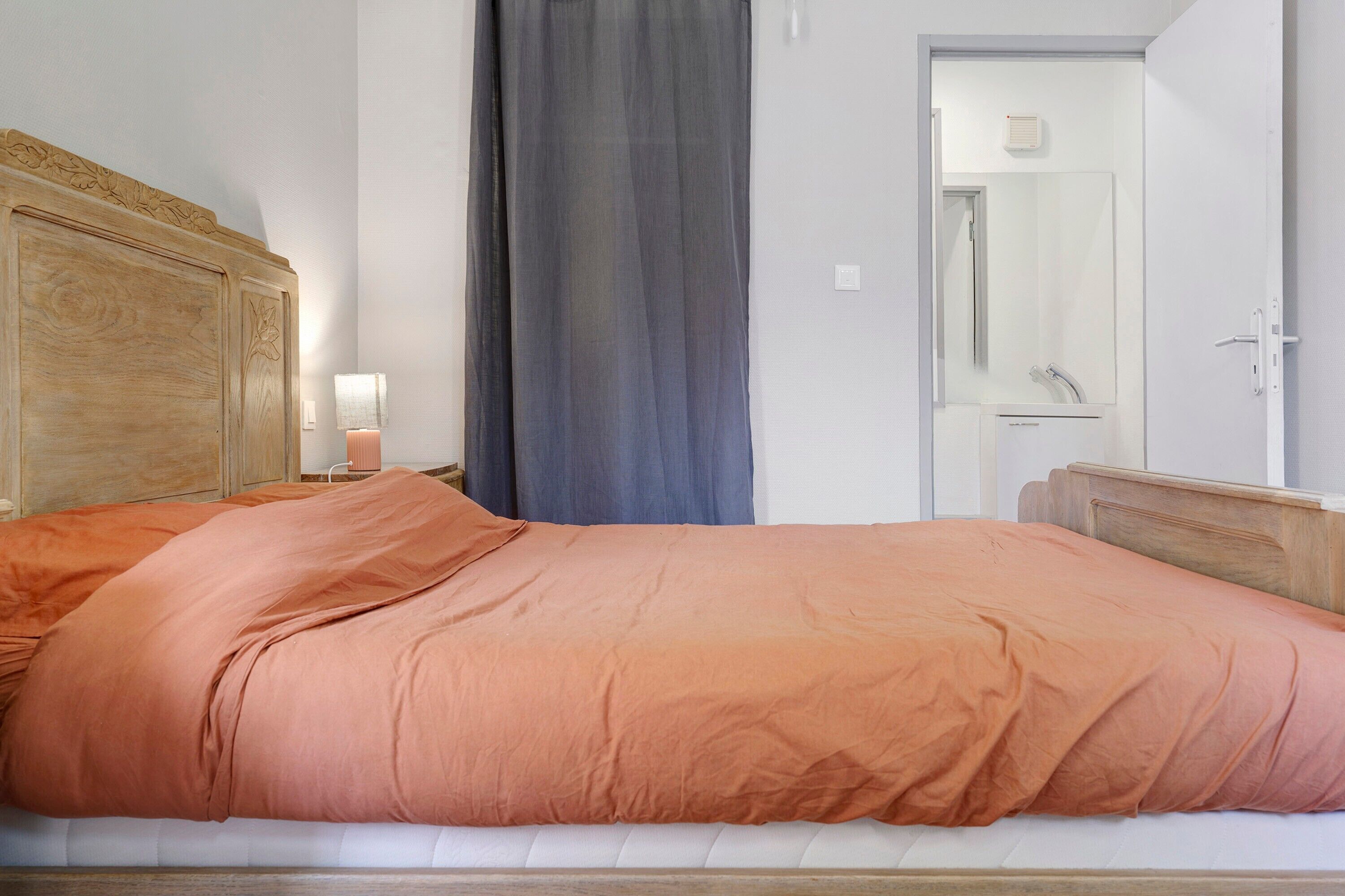 A bedroom with a wooden headboard, a bed with an orange bedspread, a nightstand with a lamp, and a bathroom door with a mirror.