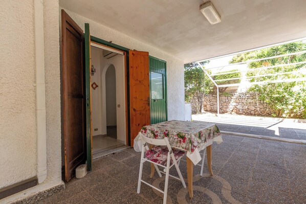 A covered outdoor seating area with a table and chairs, a wooden door, and a view of greenery and a stone wall. - Villa La Tortora - Standard by BarbarHouse (Gagliano del Capo)