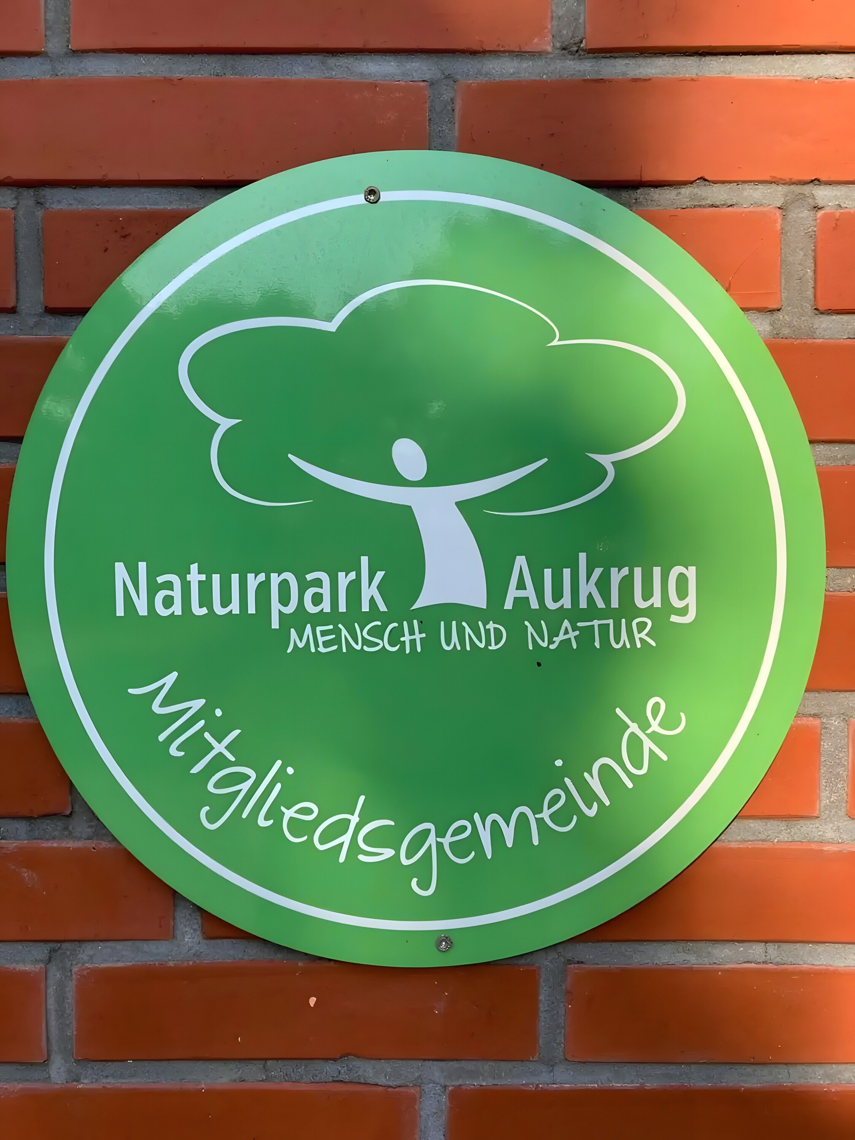 Un panneau circulaire vert, avec du texte blanc et un motif représentant un arbre, fixé sur un mur de briques.
