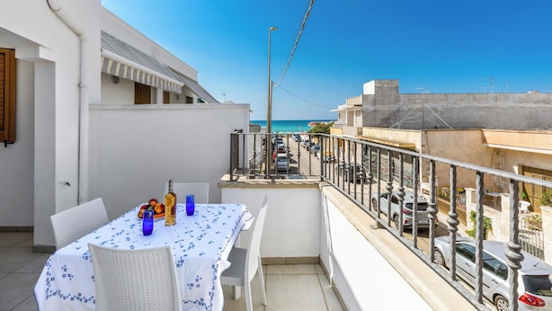 A balcony with a table set for dining, overlooking a street and the sea.