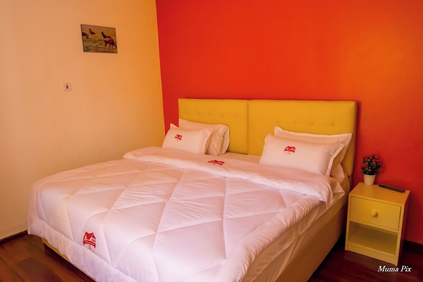 A bed with a quilted white duvet, a yellow headboard, and a small bedside table with a red logo.