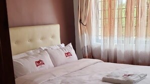 Een net opgemaakt bed met witte lakens en kussens, een beige hoofdeinde en een raam met dunne gordijnen.