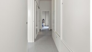 Ein schmaler Flur mit weißen Wänden und Türen, der zu einem Zimmer mit einem Bett und einer Kommode führt.