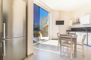 A modern kitchen with a stainless steel refrigerator, a dining table with chairs, a flat-screen TV mounted above the counter, and a staircase leading to an outdoor area.