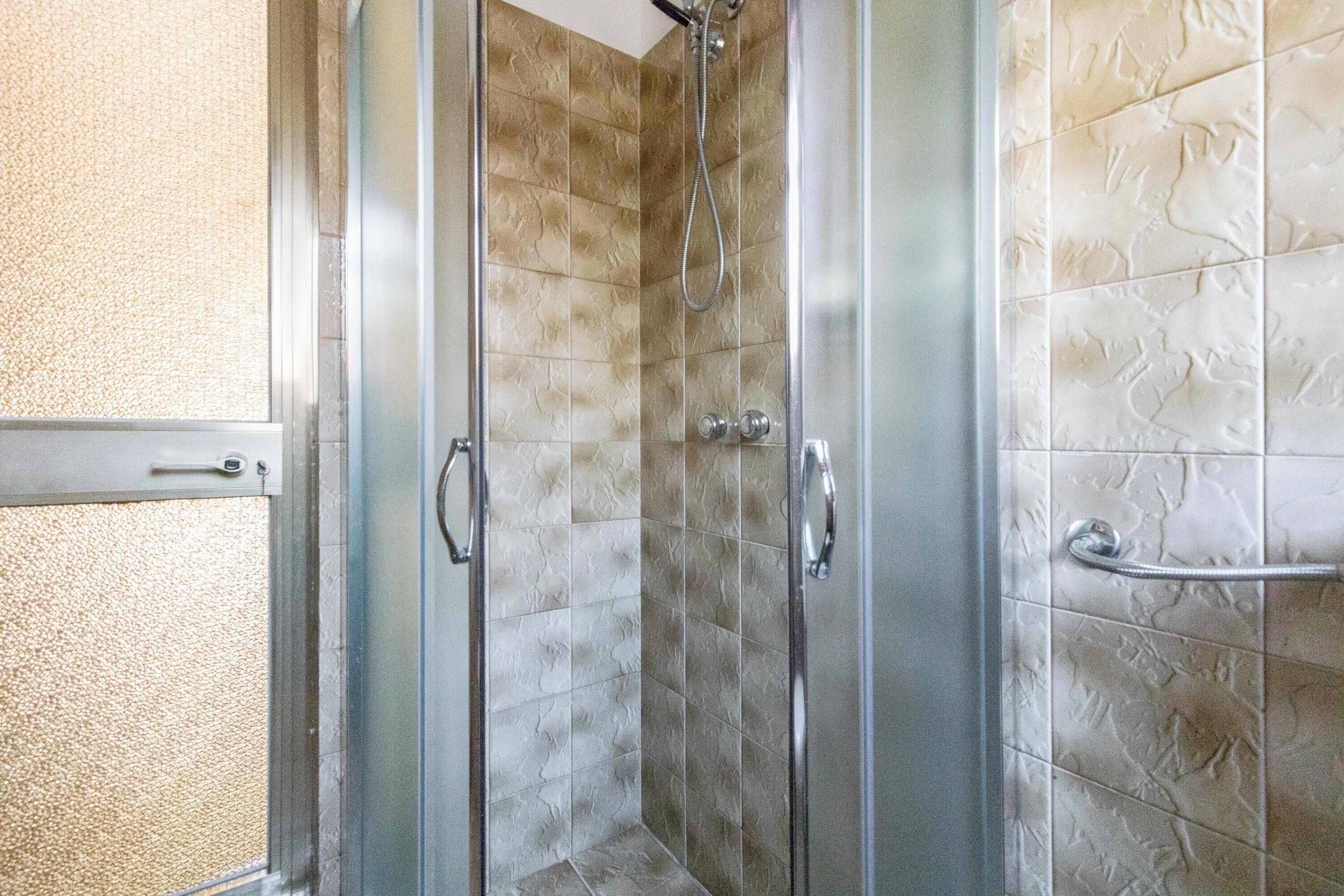 Ein Duschbereich mit Glastrennwand, einer Wand mit strukturierter Fliesenoptik und einem Metallgriff.