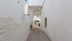 Un stretto vicolo con pareti bianche e pavimento piastrellato, che conduce a un edificio con tetto in tegole.