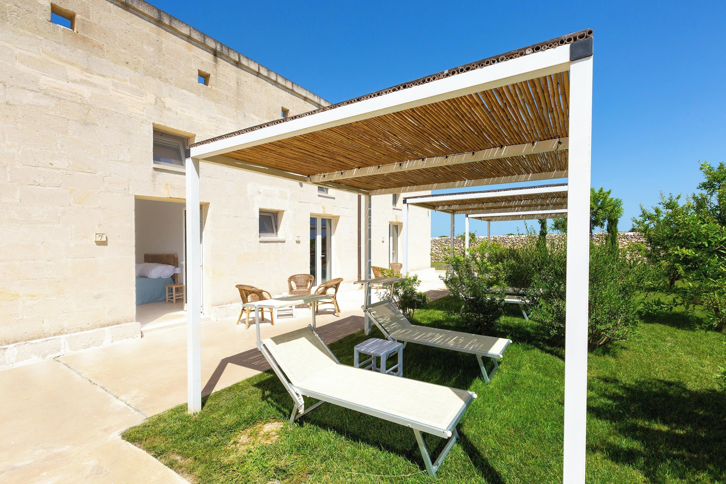 Un espace extérieur abrité avec deux chaises longues et une table, un bâtiment en pierre en arrière-plan et un ciel bleu dégagé.