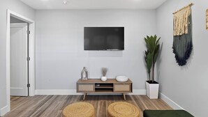 Een moderne woonkamer met een flatscreen televisie aan de muur, een houten console tafel met decoratieve objecten, een grote potplant en geweven manden op de vloer.