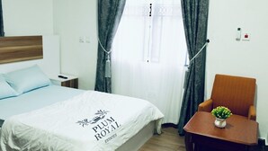 Habitación de hotel con cama, una silla, una mesita con un jarrón de flores y una ventana con cortinas.