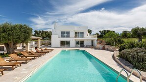 Ein modernes Haus mit zwei Etagen, einem Swimmingpool, Liegestühlen und einer Steinmauer.