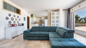Ein modernes Wohnzimmer mit einer Eckcouch, einer Küche im Hintergrund und einem großen Schiebeglasfenster zum Außenbereich.