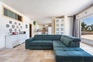 A modern living room with a sectional sofa, a kitchen in the background, and a large sliding glass door to the outside. - Villa Matilda by Perle di Puglia (Ostuni)