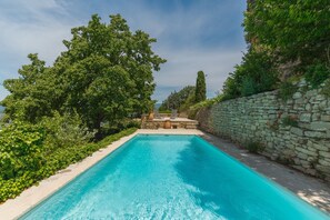A rectangular swimming pool with clear blue water, surrounded by lush greenery and a stone wall.