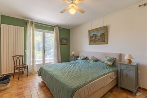 A bedroom with a bed, a nightstand, a chair, a radiator, a window with curtains, and a ceiling fan.