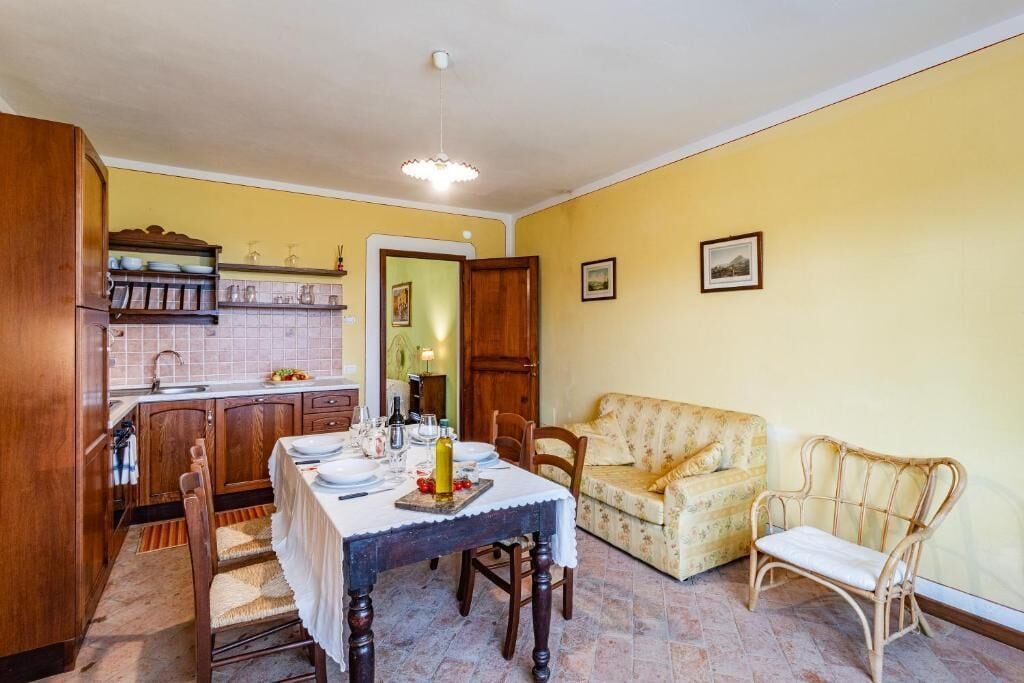 Un comedor con una mesa lista para dos personas, sillas de madera, un sofá y un sillón de mimbre. Se ve una cocina al fondo, con armarios de madera y salpicadero de azulejos.
