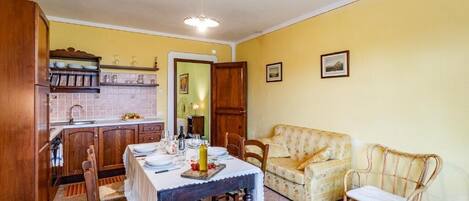 Un comedor con una mesa lista para dos personas, sillas de madera, un sofá y un sillón de mimbre. Se ve una cocina al fondo, con armarios de madera y salpicadero de azulejos.