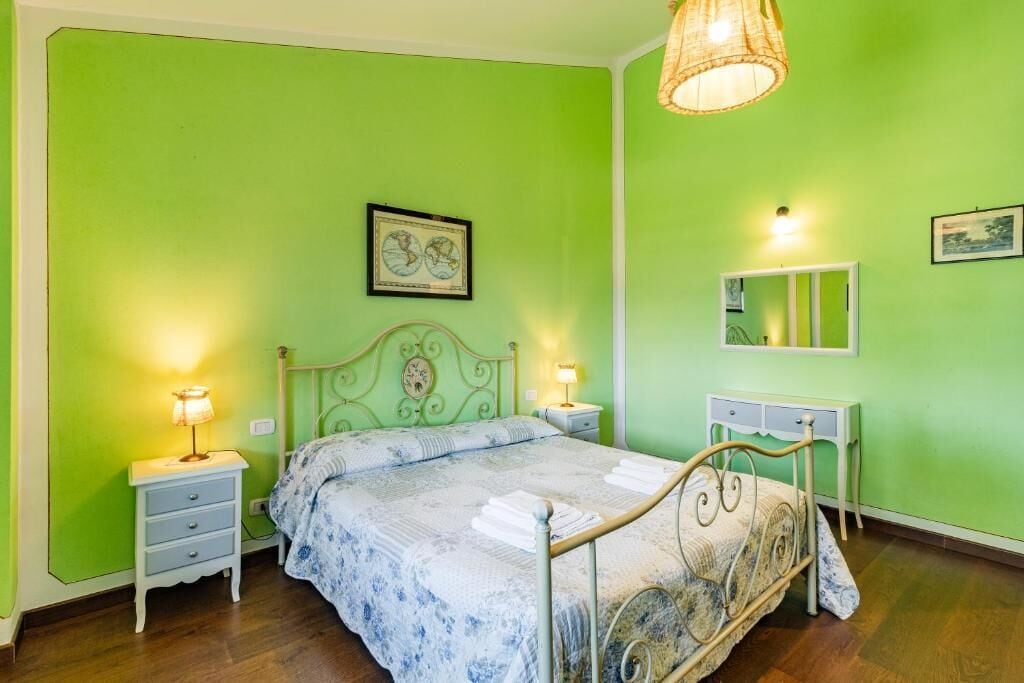 A bedroom with a green wall, a bed with a floral bedspread, bedside lamps, a nightstand, a dresser, and a hanging light fixture.
