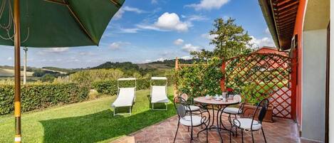 A patio with a table and chairs, a green umbrella, and a view of a grassy area and hills in the background.