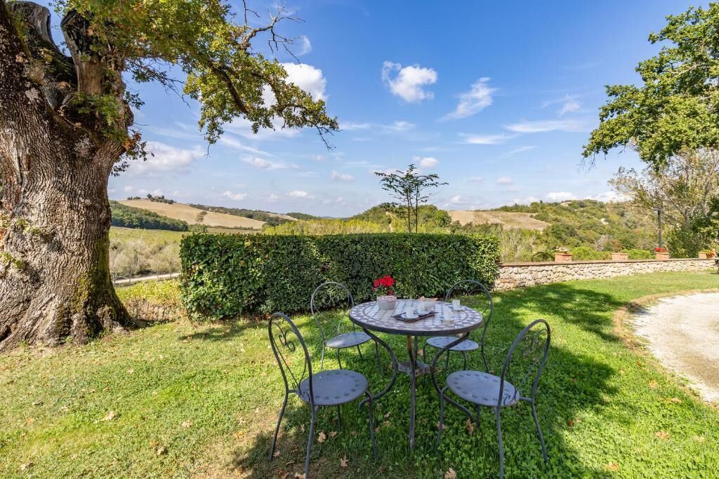 An outdoor dining area with a round metal table and chairs, surrounded by greenery and a large tree.