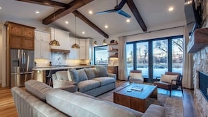 Un salon moderne comprenant un canapé, une table basse et des fauteuils, une cuisine avec des meubles en bois et un réfrigérateur, ainsi que de grandes fenêtres offrant une vue sur l’extérieur.
