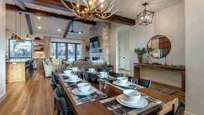 Un coin repas avec une longue table en bois dressée pour le repas, une cheminée en pierre et un grand lustre.