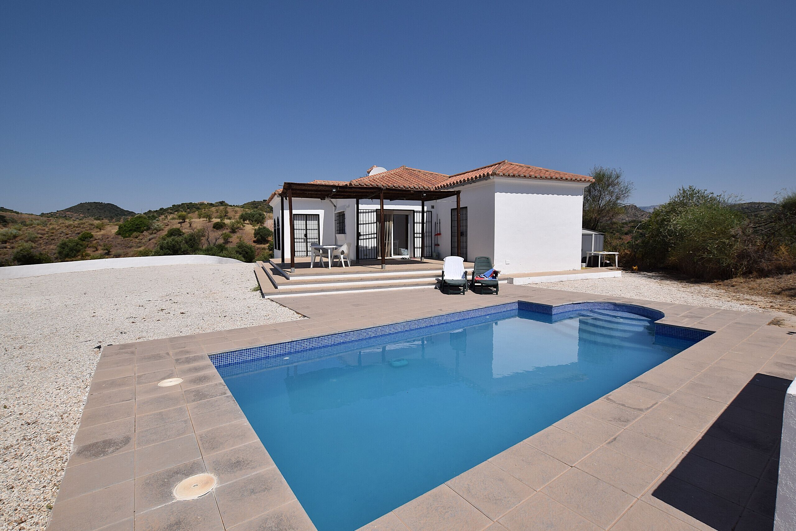 A swimming pool with a tiled deck, a covered patio with outdoor furniture, and a white building with a red roof.