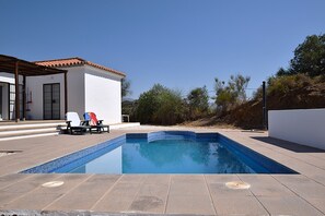 A swimming pool with a tiled edge, a covered seating area with lounge chairs, and a building with a tiled roof. - Cozy casita in the countryside with swimming pool, near Málaga (Almogía)