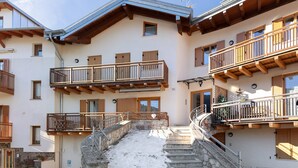 A multi-story building with balconies, a stone staircase, and a snow-covered ground. - Apartment 'Lory' with Mountain View and Balcony (Daldoss)
