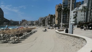 Ein Strand mit einem gepflasterten Weg, Sonnenschirmen und Gebäuden im Hintergrund.