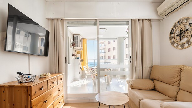 Ein modernes Wohnzimmer mit einer Couch, einem Fernseher, einer Holzkommode und Blick durch eine Glastür auf einen Außenbalkon.