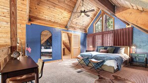 A rustic bedroom with a wooden ceiling, a bed with blue bedding, a desk with a clock, and a leather rug. - Rushmore Retreat - View of Mt Rushmore + Hot Tub + Game Room (Keystone)