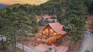 A log cabin with a deck and outdoor seating, surrounded by a forest. - Rushmore Retreat - View of Mt Rushmore + Hot Tub + Game Room (Keystone)
