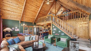 A spacious living area with a wooden ceiling, a green couch, a brown sofa, a dining table, and a staircase leading to an upper level. - Rushmore Retreat - View of Mt Rushmore + Hot Tub + Game Room (Keystone)