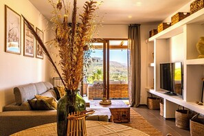 Ein modernes Wohnzimmer mit einer Couch, einem Couchtisch, einer Vase mit Trockenpflanzen, einem Fernseher auf einem Regal und Blick auf eine gebirgige Landschaft durch eine Glastür.