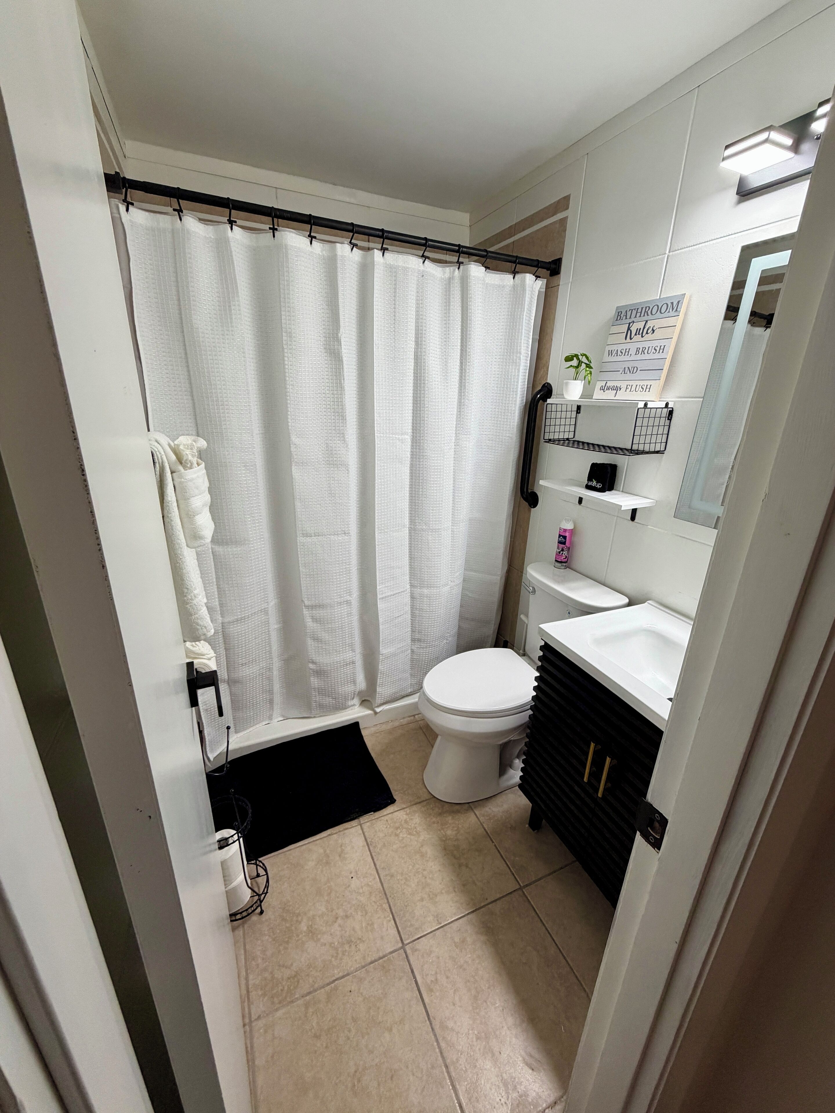 A small, clean bathroom with a toilet, sink, and shower curtain. There is a sign on the wall and a small shelf with toiletries.